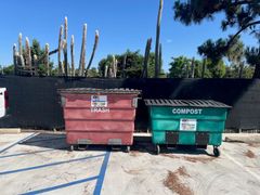 Red trash bin and green compost bin in parking lot, with fence and cactus in background.