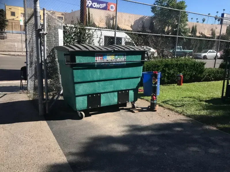 Green dumpster near a chain-link fence, parked on pavement next to grass. A sign is visible in the background.