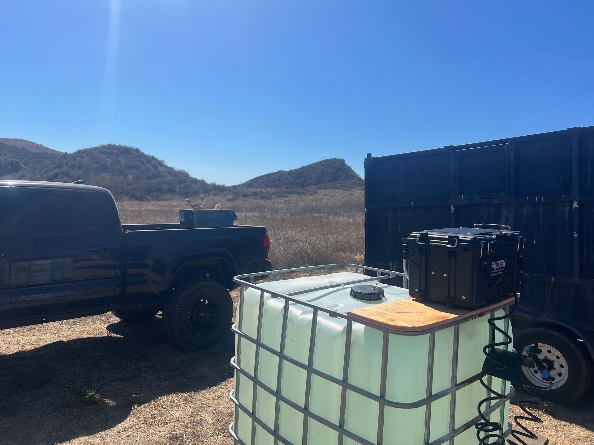 A black pickup truck, a water tank, and a trailer parked outdoors on a sunny day.