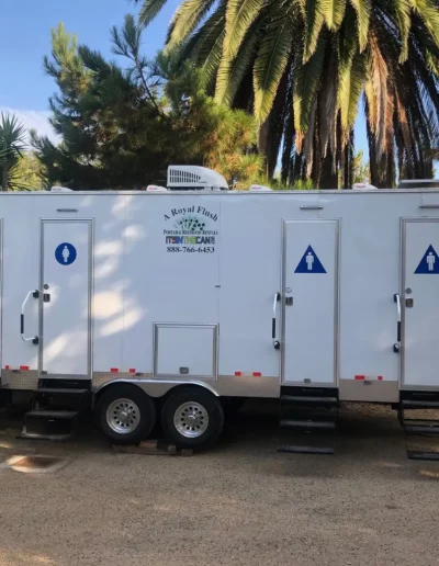 White portable restroom trailer with three individual stalls; outdoors, daytime.