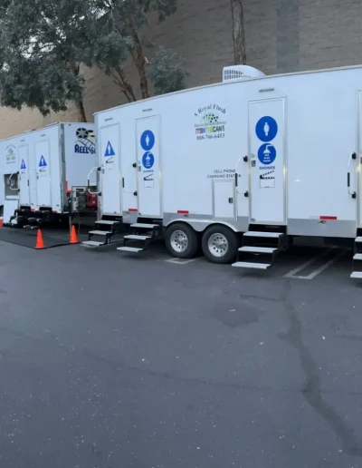 Portable restrooms on trailers. White with blue gender signs. Set on pavement next to a building.