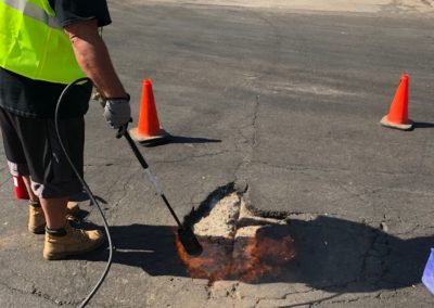 Worker using a torch to heat asphalt to repair a pothole. Wearing a yellow vest, orange cones visible.
