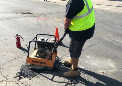 Person in safety vest using a plate compactor to repair asphalt on a road.
