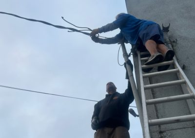 Two people working on electrical wires near a building using a ladder. Overcast sky.
