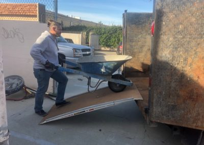 Person loading a wheelbarrow into a dumpster on a ramp. Outdoors, sunny day.