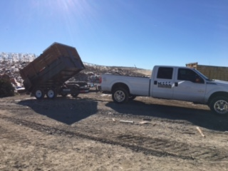 White pickup truck towing a dump trailer filled with debris against a sunny, blue sky.