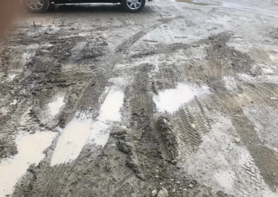 Muddy, wet dirt road with tire tracks and standing water; car in the background.
