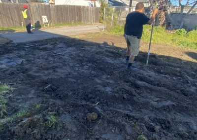 Two people preparing a patch of dirt for planting in a yard. One holds a pole, the other stands.