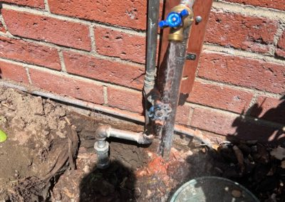 Water pipes and spigot next to a brick wall. A small hole in the ground reveals some pipe fittings.