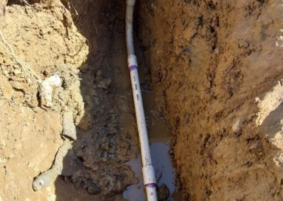 Trench with white PVC pipe and fittings, possibly for a water system, in brown soil.