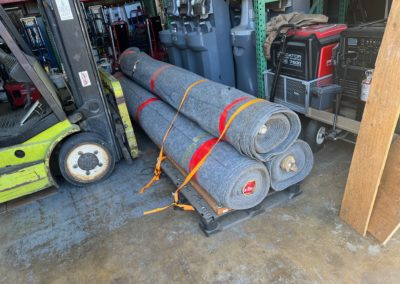 Pallet with three rolled-up gray carpets, secured with straps, next to a forklift in a warehouse.