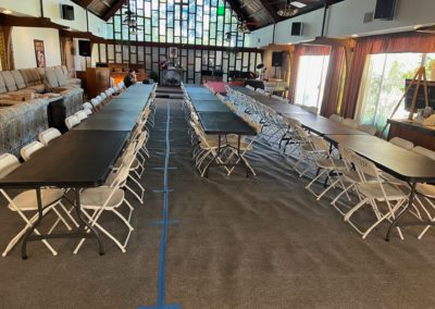 Long tables with white chairs arranged in rows inside a building with stage and stained glass windows.