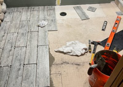 Shower floor tiling in progress: gray wood-look tiles are being laid with tools like a level and bucket of mortar.