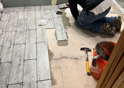 Person tiling a bathroom floor with gray wood-look tiles. Tools and bucket visible.