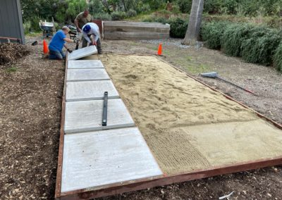 Three people laying paving stones in a yard, a level sits on one. Sand is spread in the prepared area.