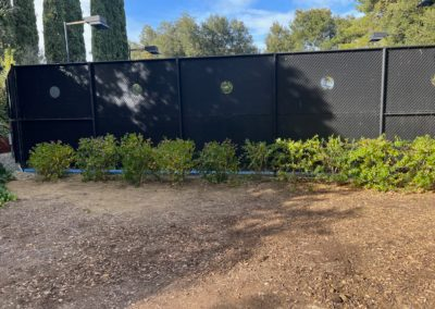 Black fence with circular accents behind a row of green bushes, under a sunny sky.