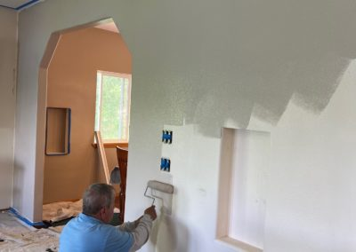 Person painting a white wall with a roller. Interior shot shows an archway and electrical boxes.