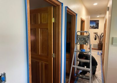 Person painting doorway, blue tape visible. Wooden door and frames contrast with white walls. Hallway setting.