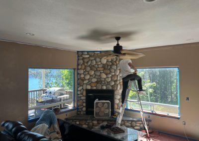 Two people painting a room, one on a ladder near a stone fireplace, windows showing a lake view.