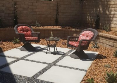Two wicker chairs with red cushions on a patio, small table, rock border, and a raised brick planter.