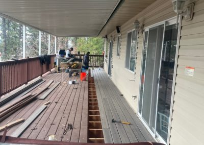 Deck replacement in progress; boards removed. Two workers, tools, and house with sliding door visible.