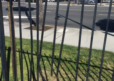 Black metal fence casting shadows on green grass and sidewalk near a street.