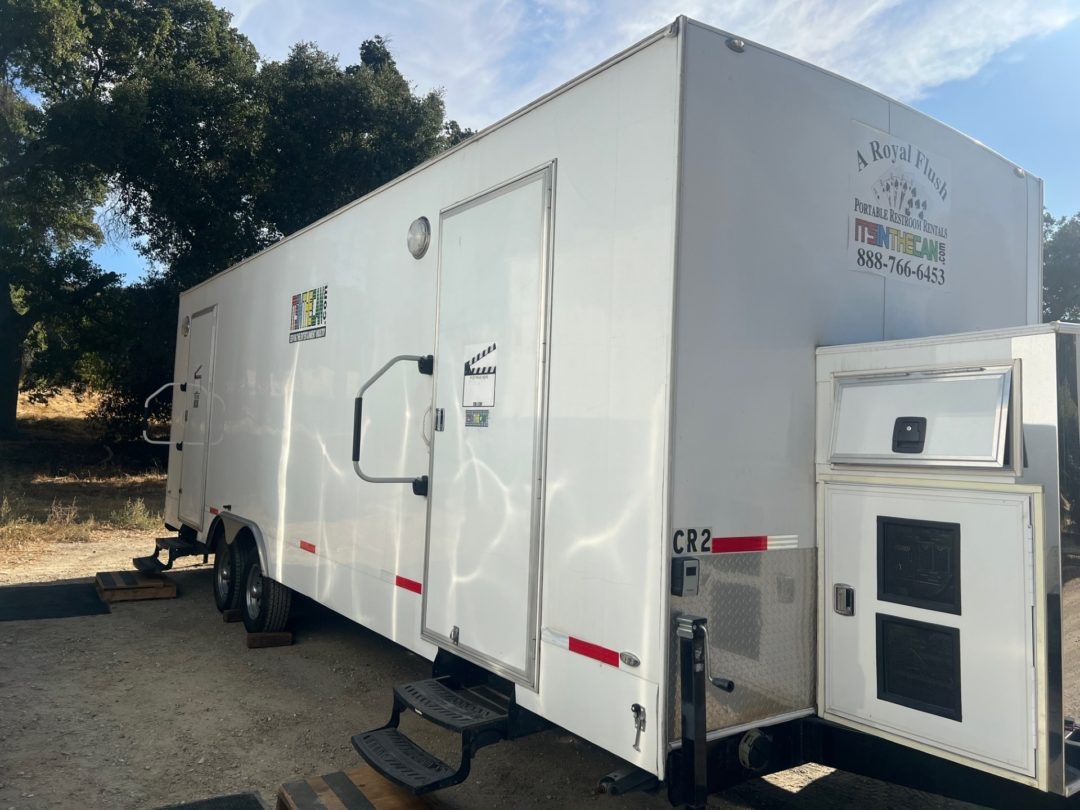 White portable restroom trailer on a gravel surface outdoors, with a service door open.