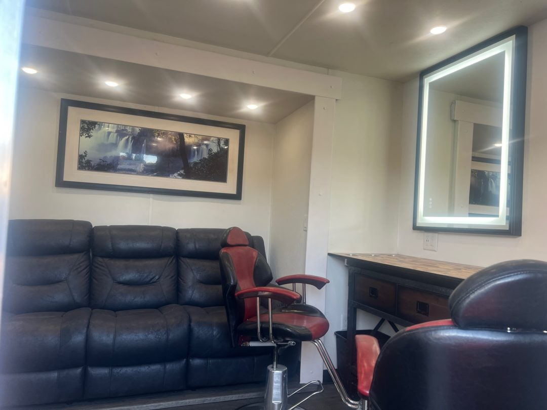 Barbershop interior with a black leather couch, red and black barber chair, framed art, and a lighted mirror.