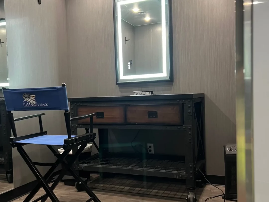 Makeup vanity area with lit mirror, dark wood table, director's chair. Gray walls.