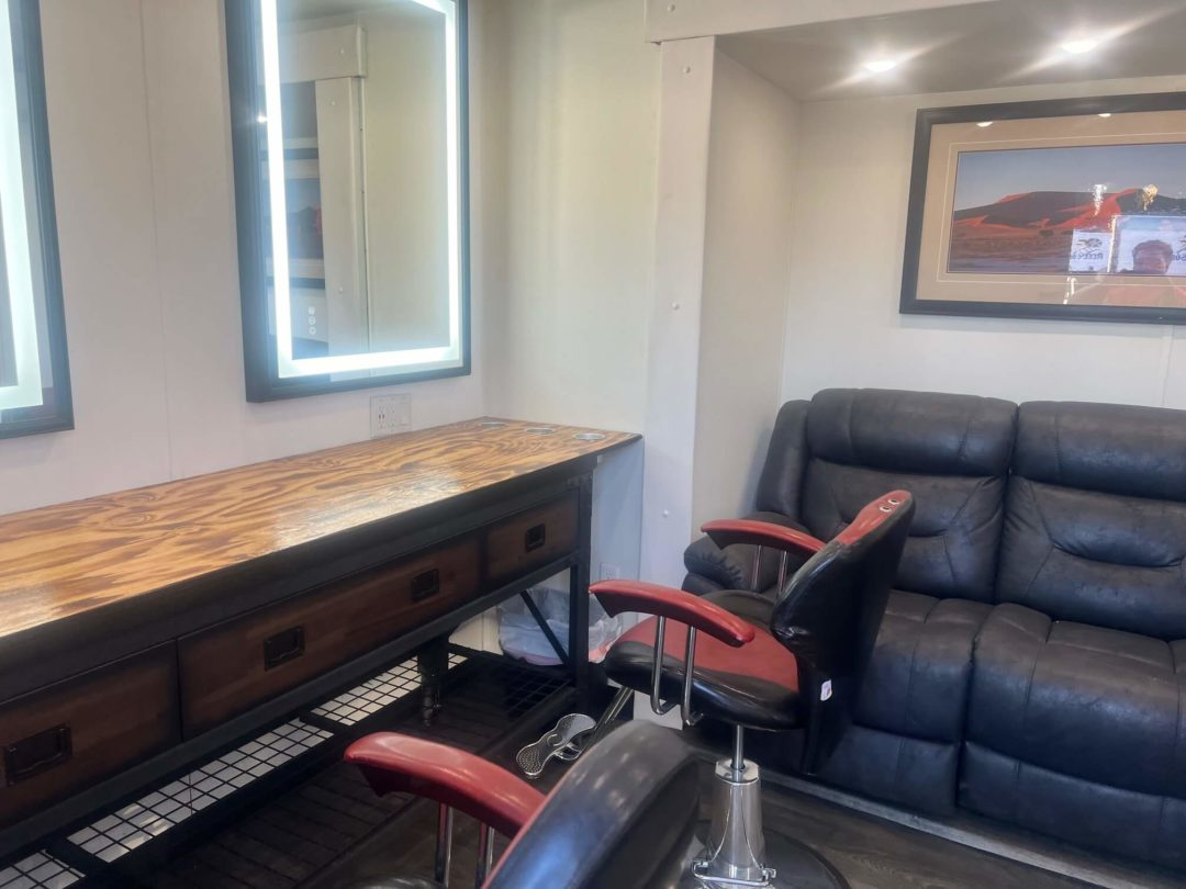 Salon interior with a workstation, mirror, barber chair, and a waiting area with a couch and artwork.
