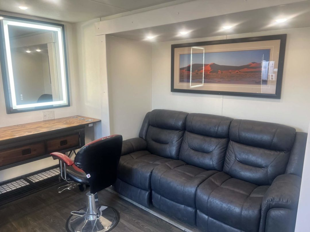 Hair salon interior: lighted mirror, seating area with black sofa and art, and a styling chair.