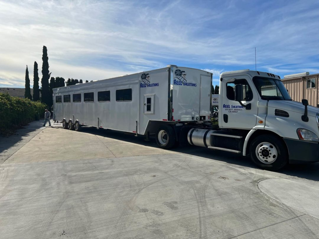 Silver horse trailer hitched to a white truck on a paved surface, under a partly cloudy sky.