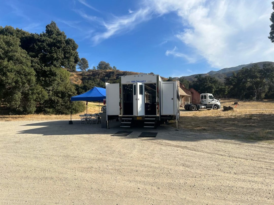 White trailer with open doors on a gravel lot under a blue sky, trees in the background.