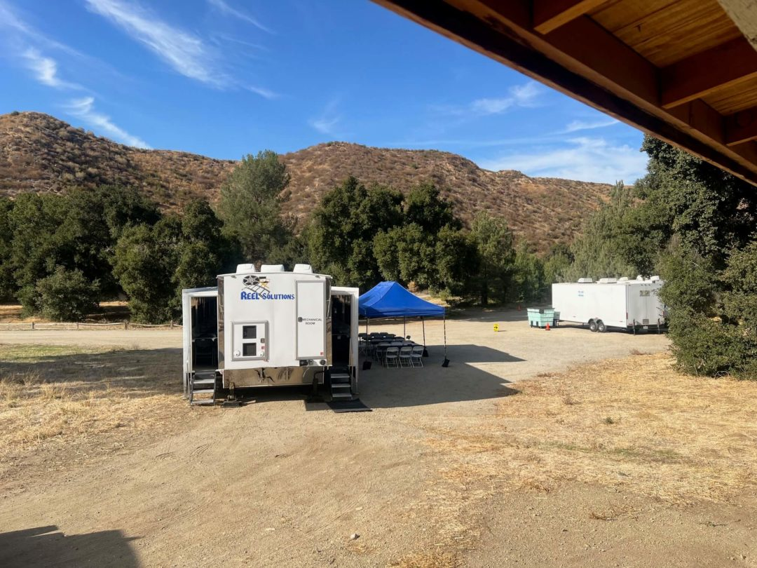 Trailers and blue tent in a dusty outdoor setting, with trees and a mountain in the background.