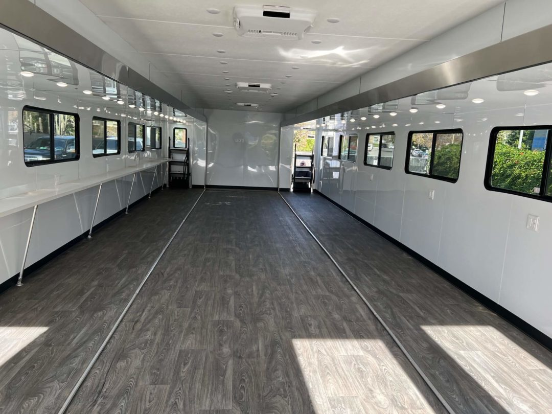 Interior of a trailer, empty with dark gray flooring, white walls, small windows, and overhead lights.