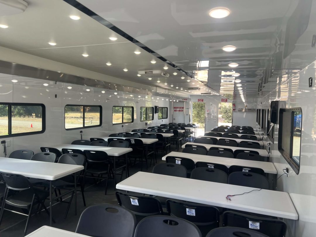 Interior of a white trailer, tables with black chairs arranged for a large group.