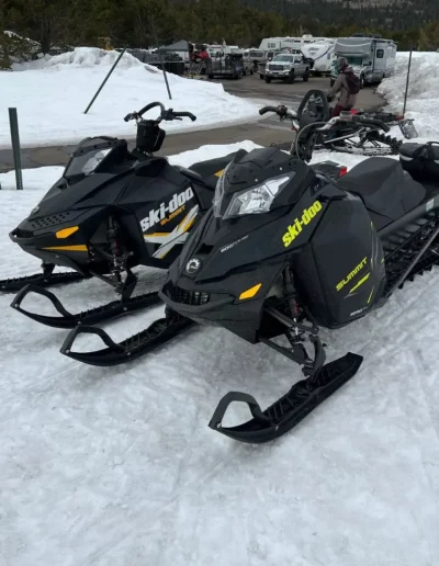 Two black and yellow Ski-Doo snowmobiles parked on snow, other vehicles in background.