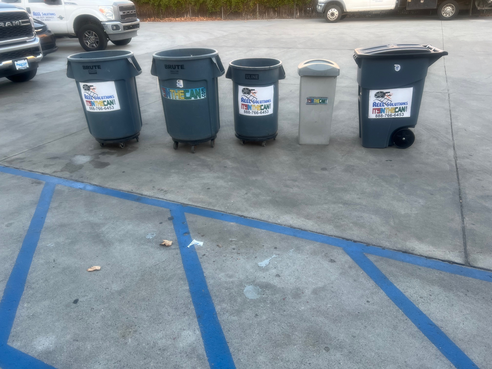 Five different trash cans of varying sizes on a gray concrete surface, with a truck and golf cart in the background.