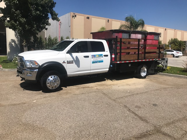 White work truck with a flatbed carrying a dumpster, parked in a lot, sunny day.