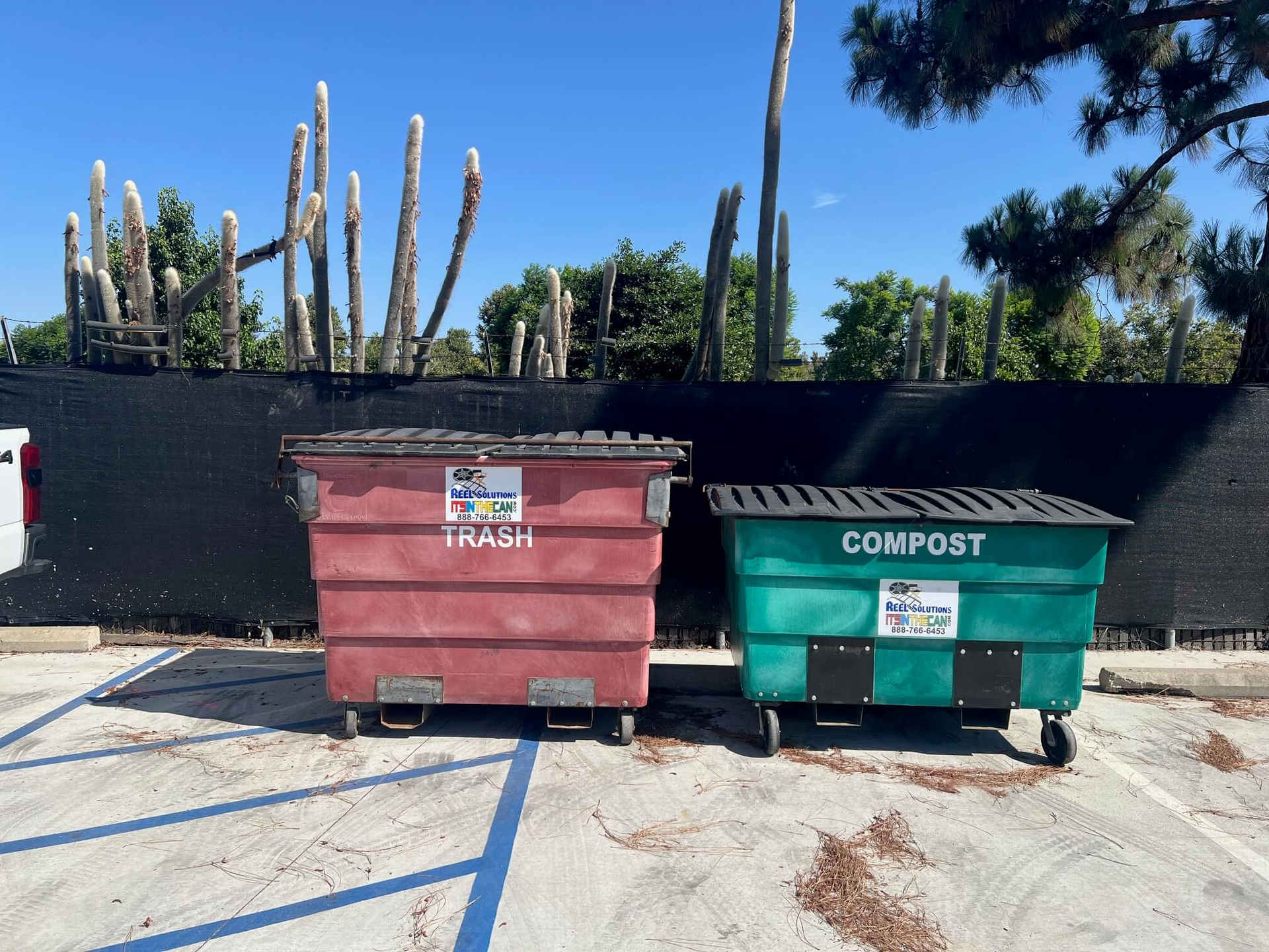 Red trash bin and green compost bin in a parking lot, with a black fence and cacti in the background.