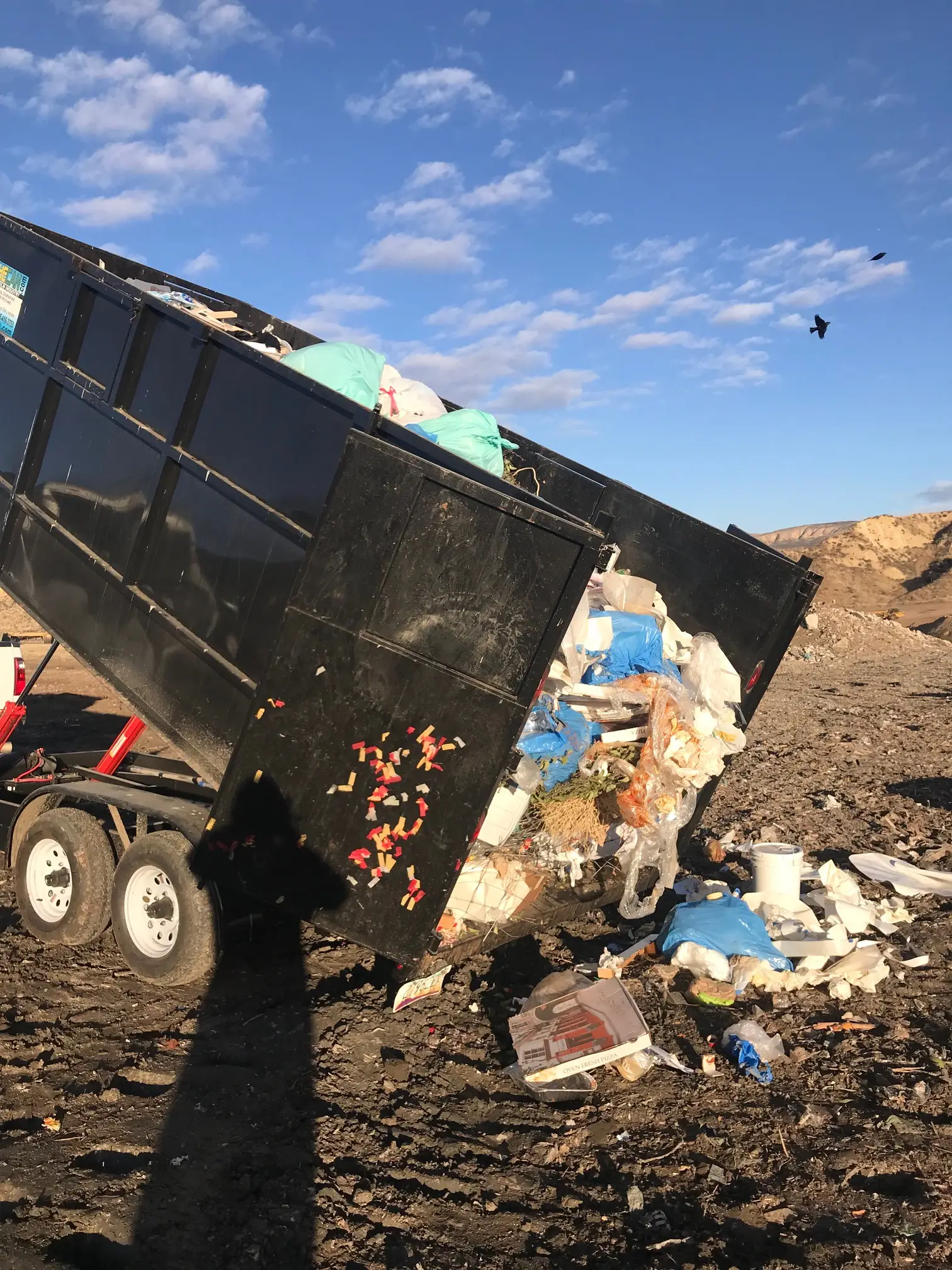 Dump truck emptying trash onto a landfill under a blue sky.
