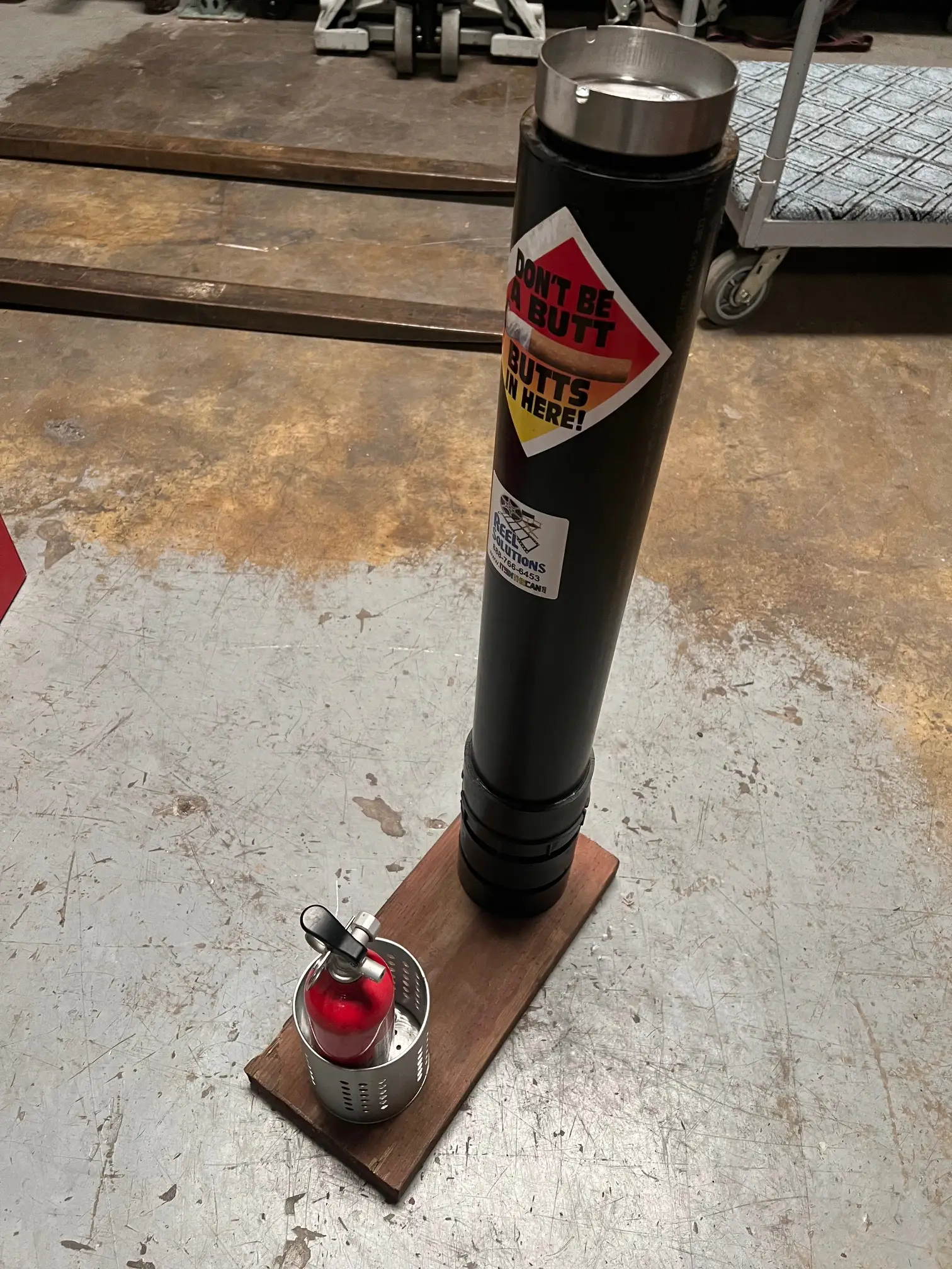 Black cylindrical object with warning label, mounted on wood base, next to fire extinguisher. Industrial setting.