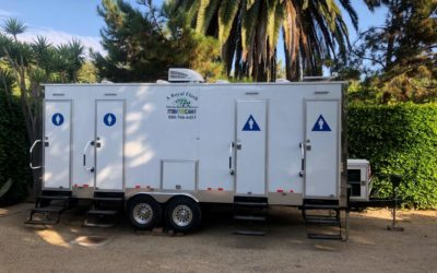 White portable restroom trailer with four stalls, in a yard with trees.