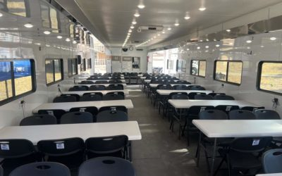 Interior of a large trailer converted into a classroom with tables, chairs, and windows.