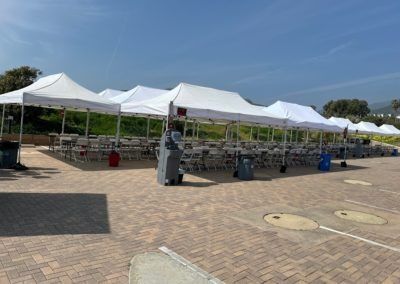 Several white tents with tables and chairs set up in a parking lot on a sunny day.