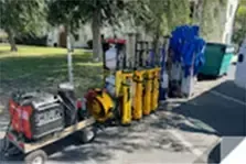 A trailer with a red generator and various yellow and blue utility items parked on pavement near green bins and trees.
