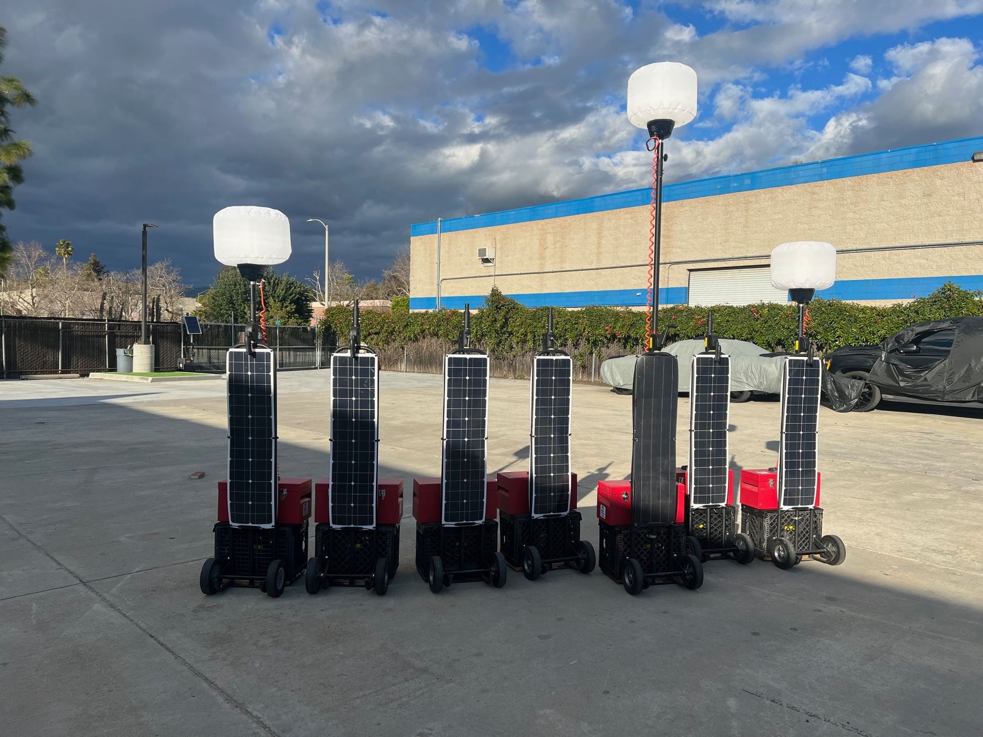 Several solar-powered mobile light towers in a row; red bases, black solar panels, and white light globes.