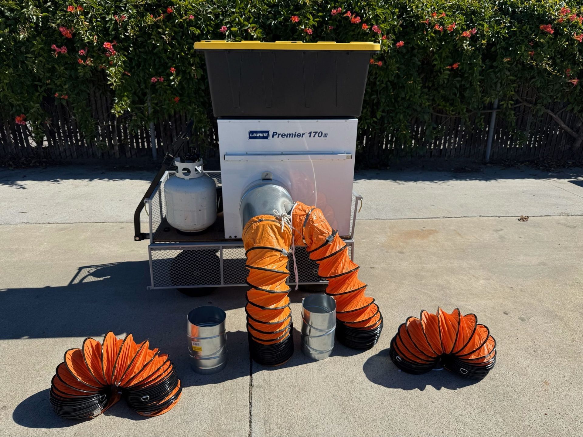 A portable propane smoker with orange vent hoses, a gray propane tank, and a black container on a wheeled cart, outdoors.
