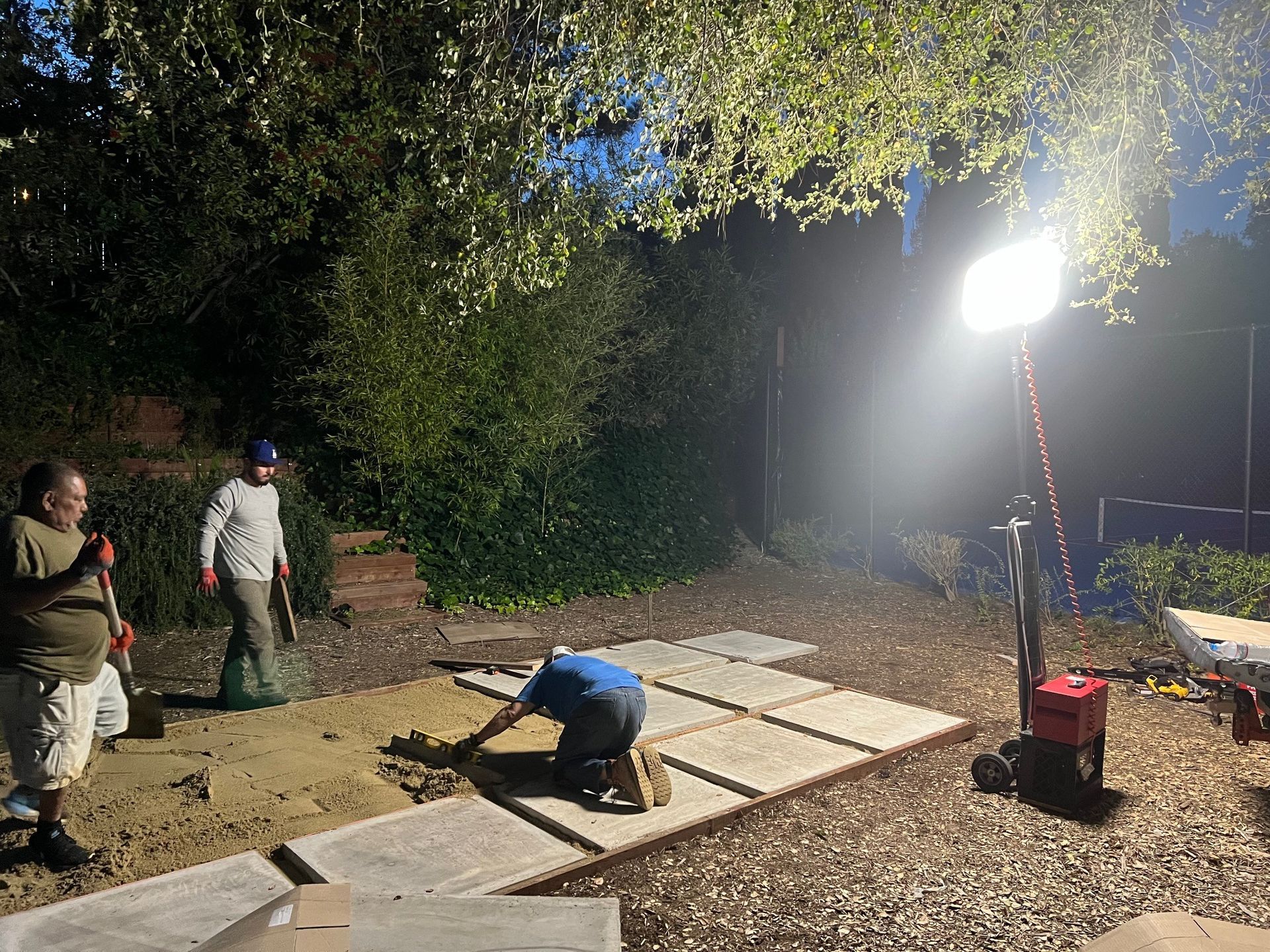 People laying pavers outdoors at night, illuminated by a work light.