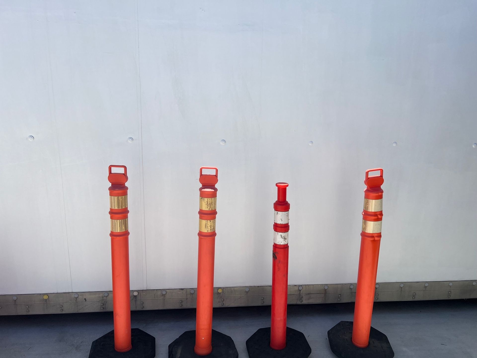 Four orange and white traffic delineator posts with black bases against a white wall.